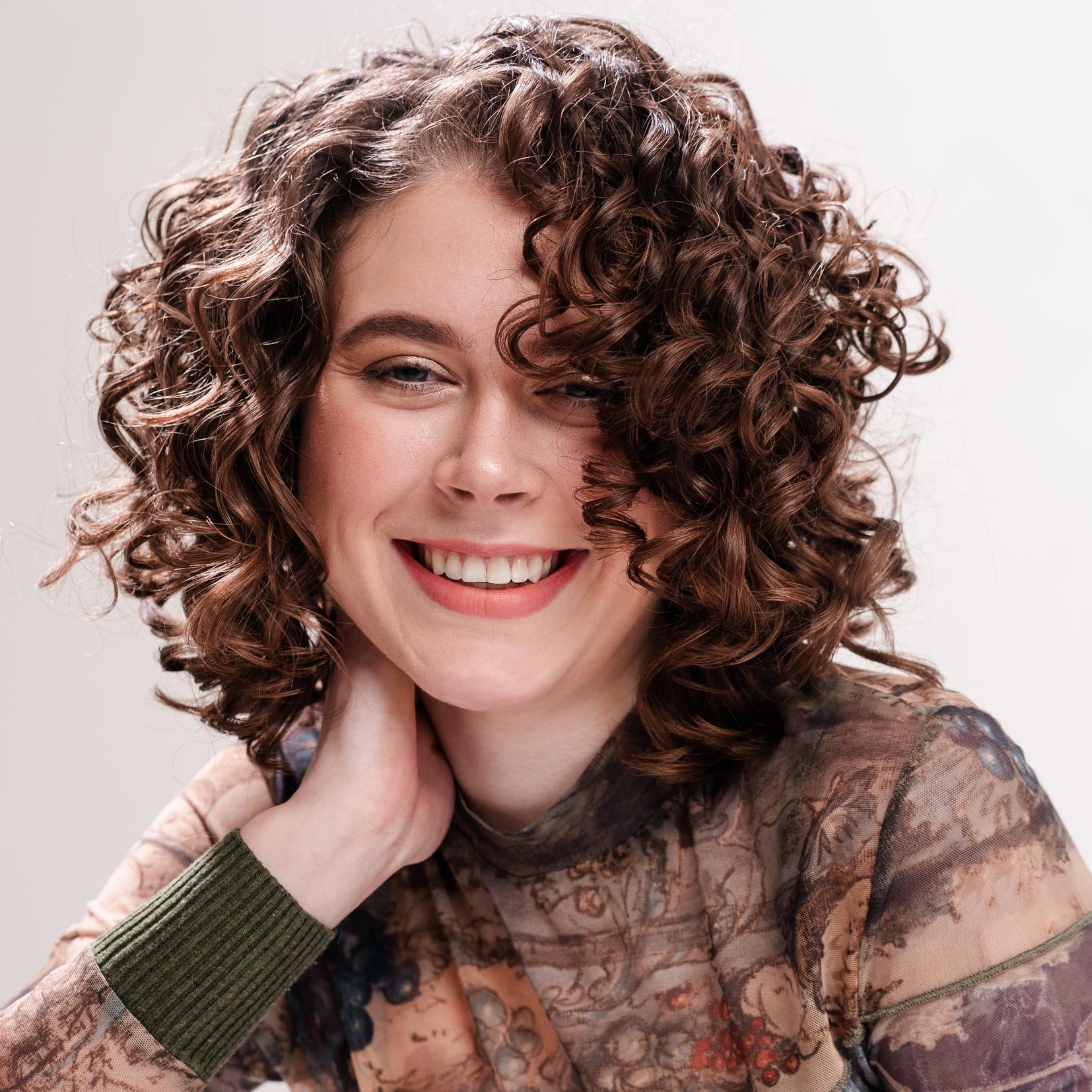 Frau mit kurzen, braunen Locken l&auml;chelt entspannt in die Kamera