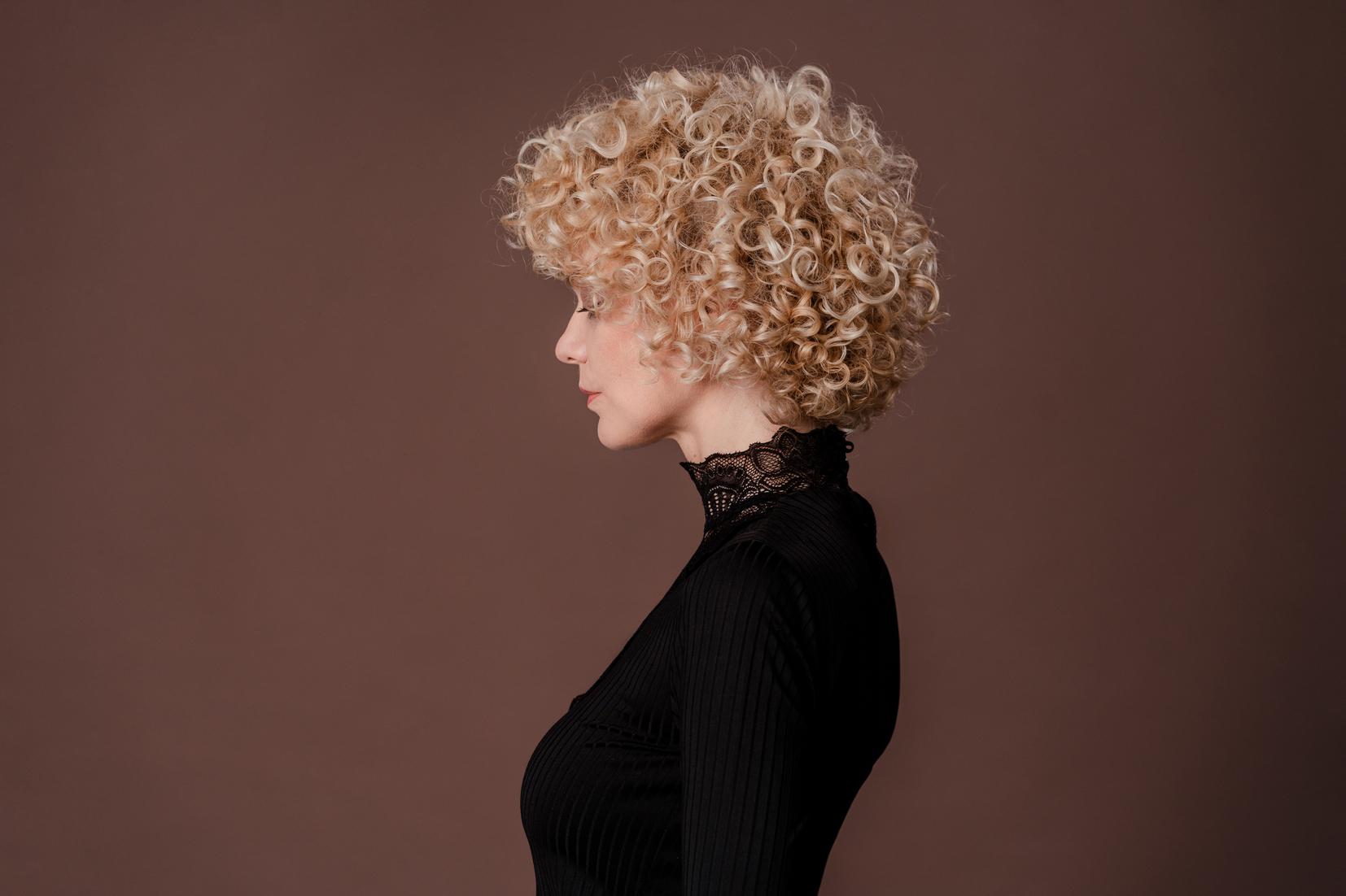 Seitenprofil einer Frau mit kurzem, blondem Lockenschnitt vor warmem Hintergrund