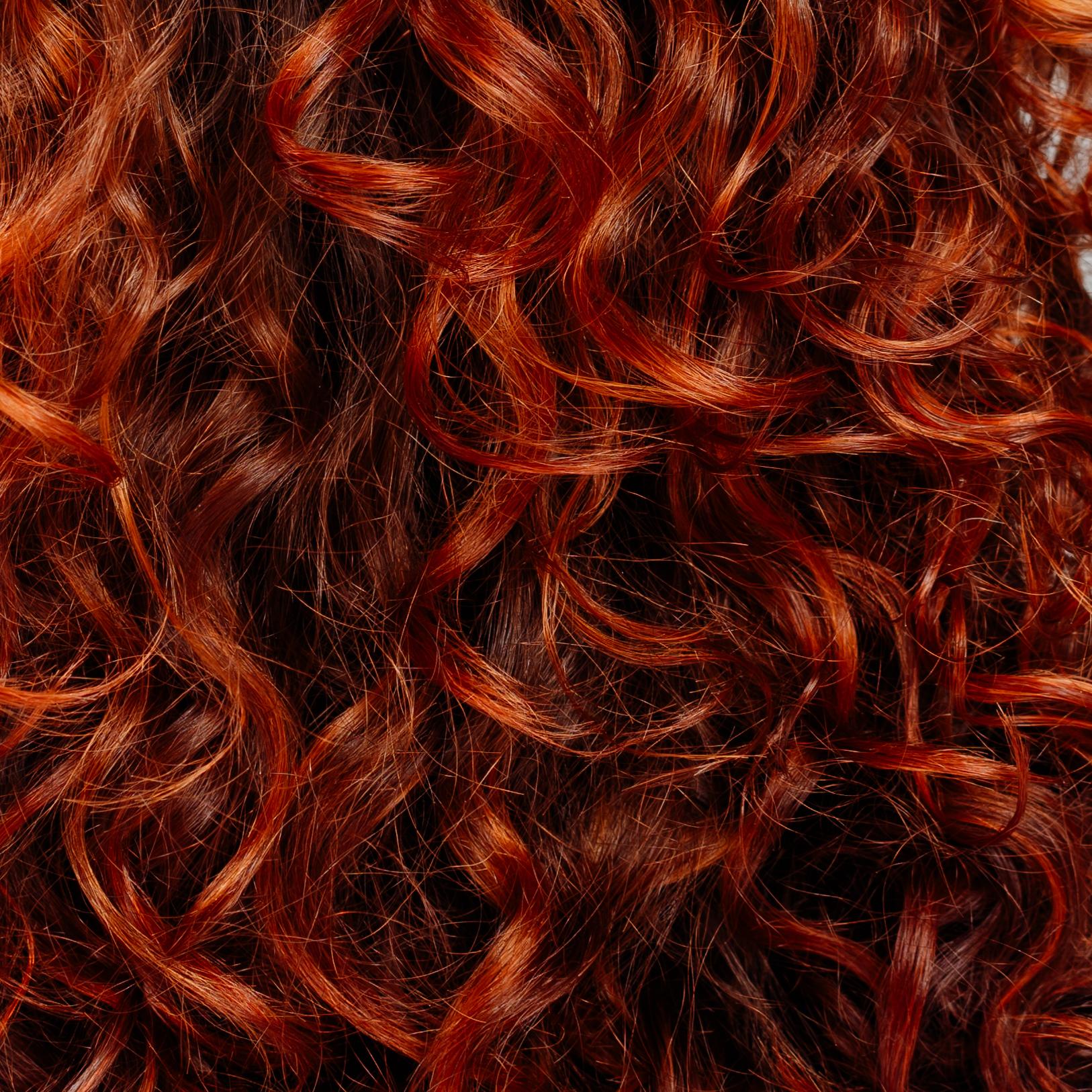 Rückansicht einer Person mit lockigem, rotbraunem Haar vor neutralem Hintergrund.