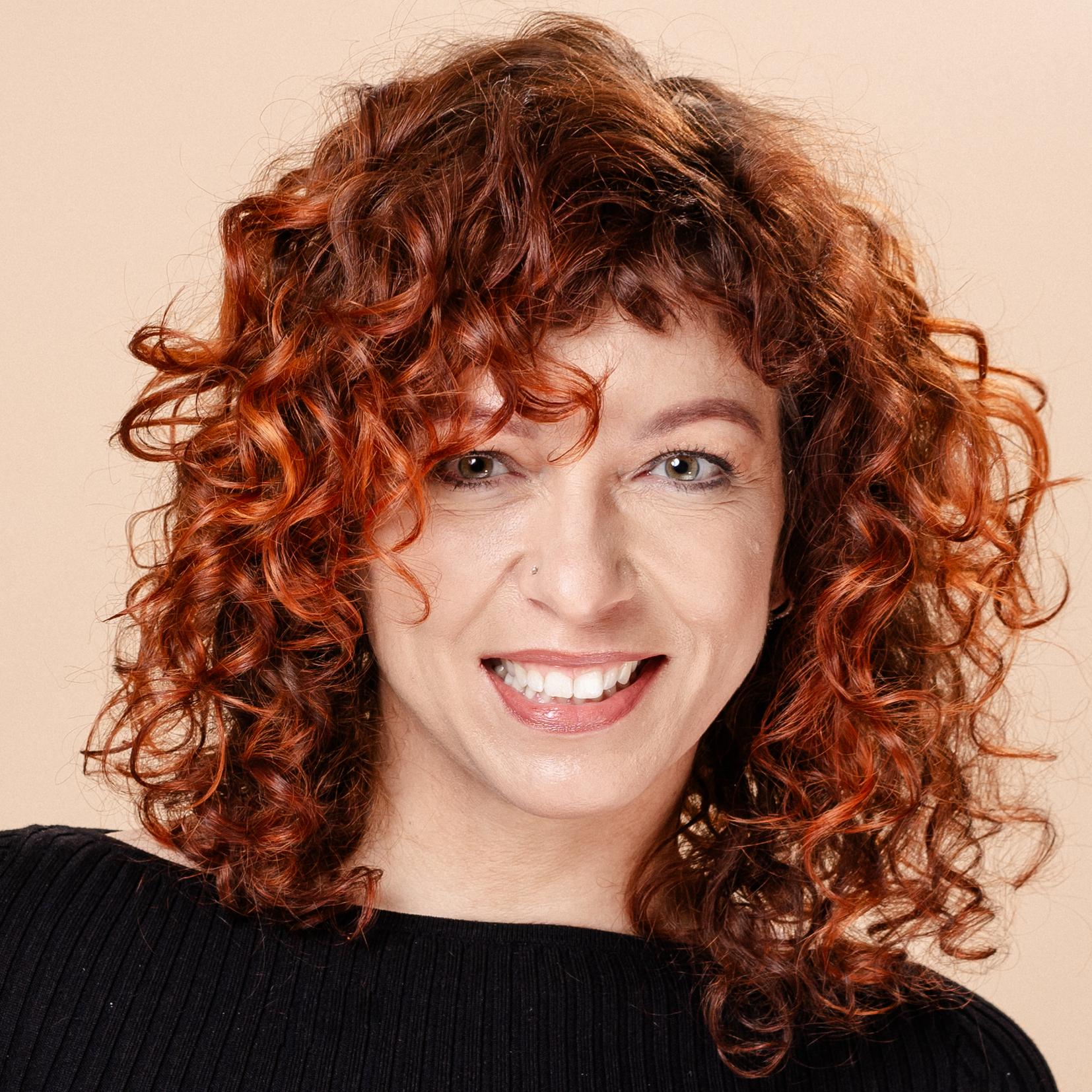 Frau mit lockigem, schulterlangem Haar in schwarzem Oberteil vor beige Hintergrund, lächelt und steht mit einer Hand an der Hüfte.