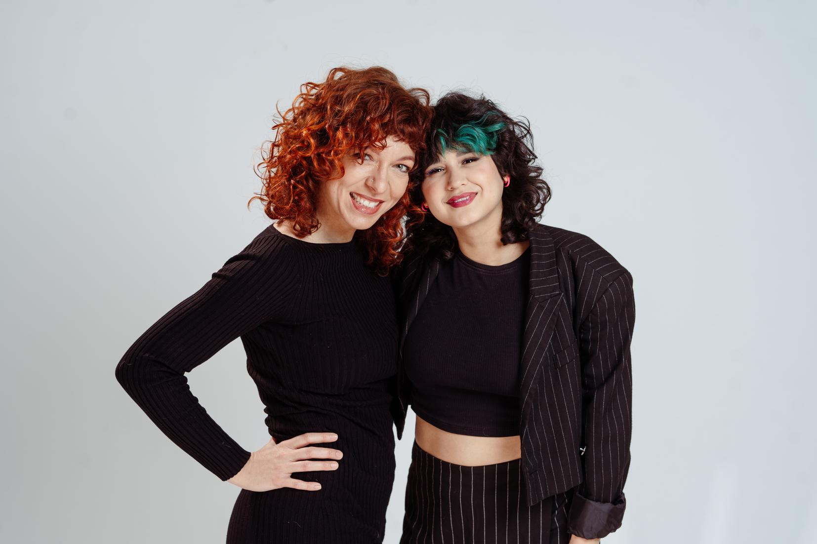 Zwei Frauen mit lockigem, rotem und schwarzem Haar l&auml;cheln zusammen vor einem hellen Hintergrund. Beide tragen schwarze Outfits.